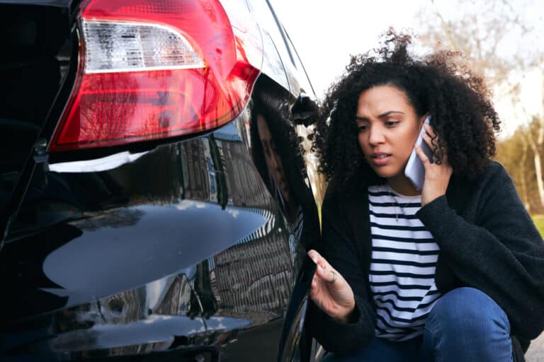A woman looking at the damaged bumper of her car after a hit and run accident, wondering what to do.